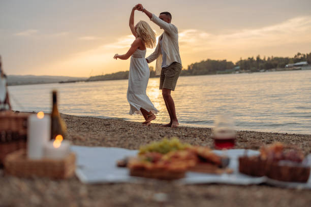 Interracial couple in love is by the river side, happily dancing the evening away at sunset after enjoying a delightful picnic.
